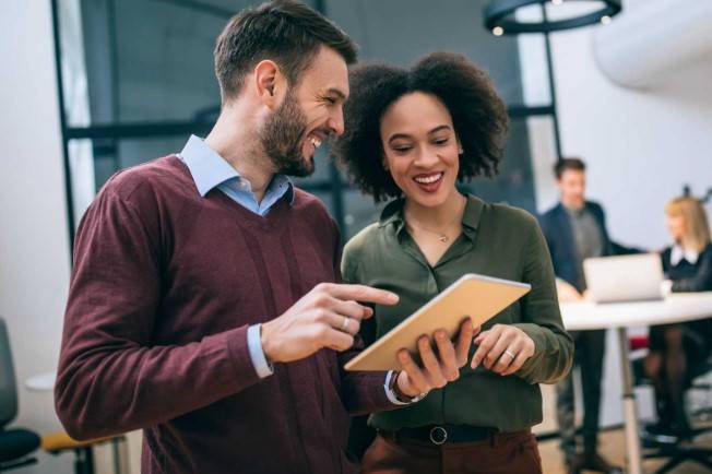 A picture of two people at a workplace looking at an ipad and smiling