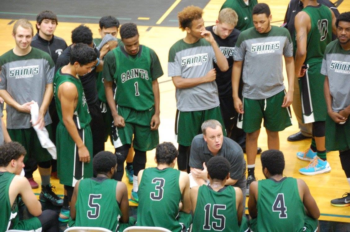 A coach kneels on the sideline of a basketball game, talking to his players