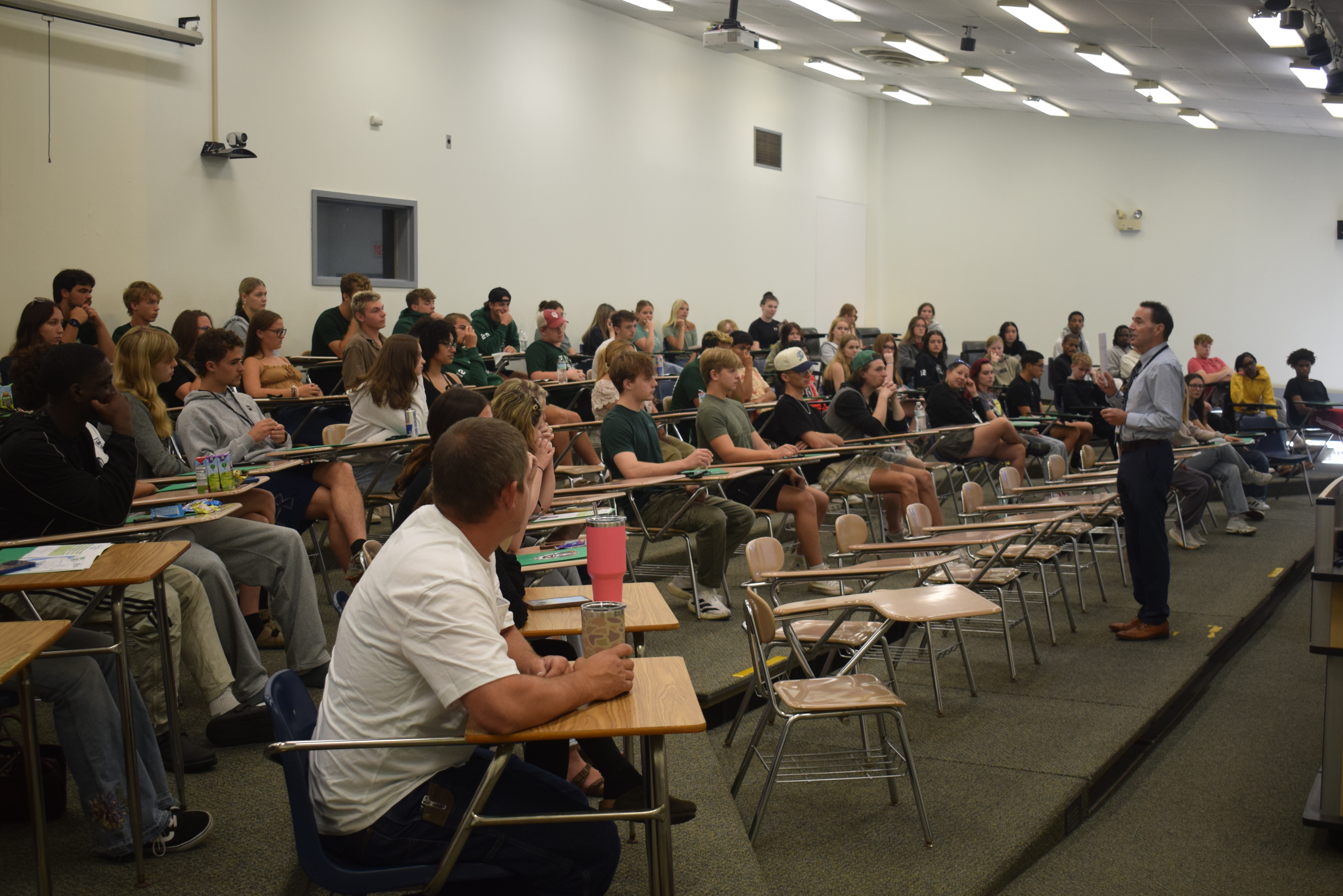 A college president speaks to a large group of students in a lecture hall