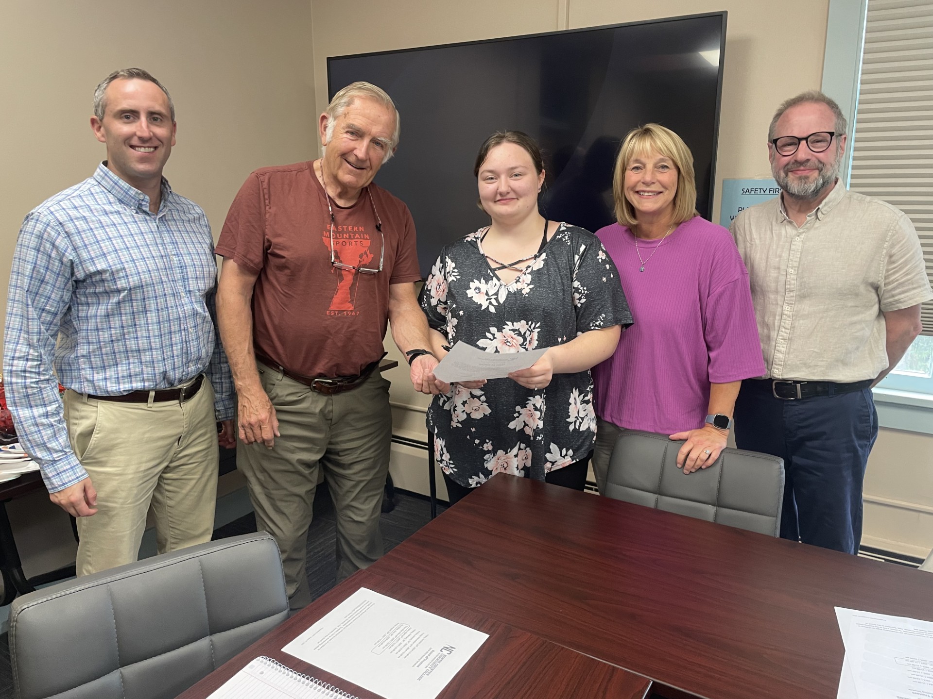 A group of people stand together and smile at the camera in a college board meeting room