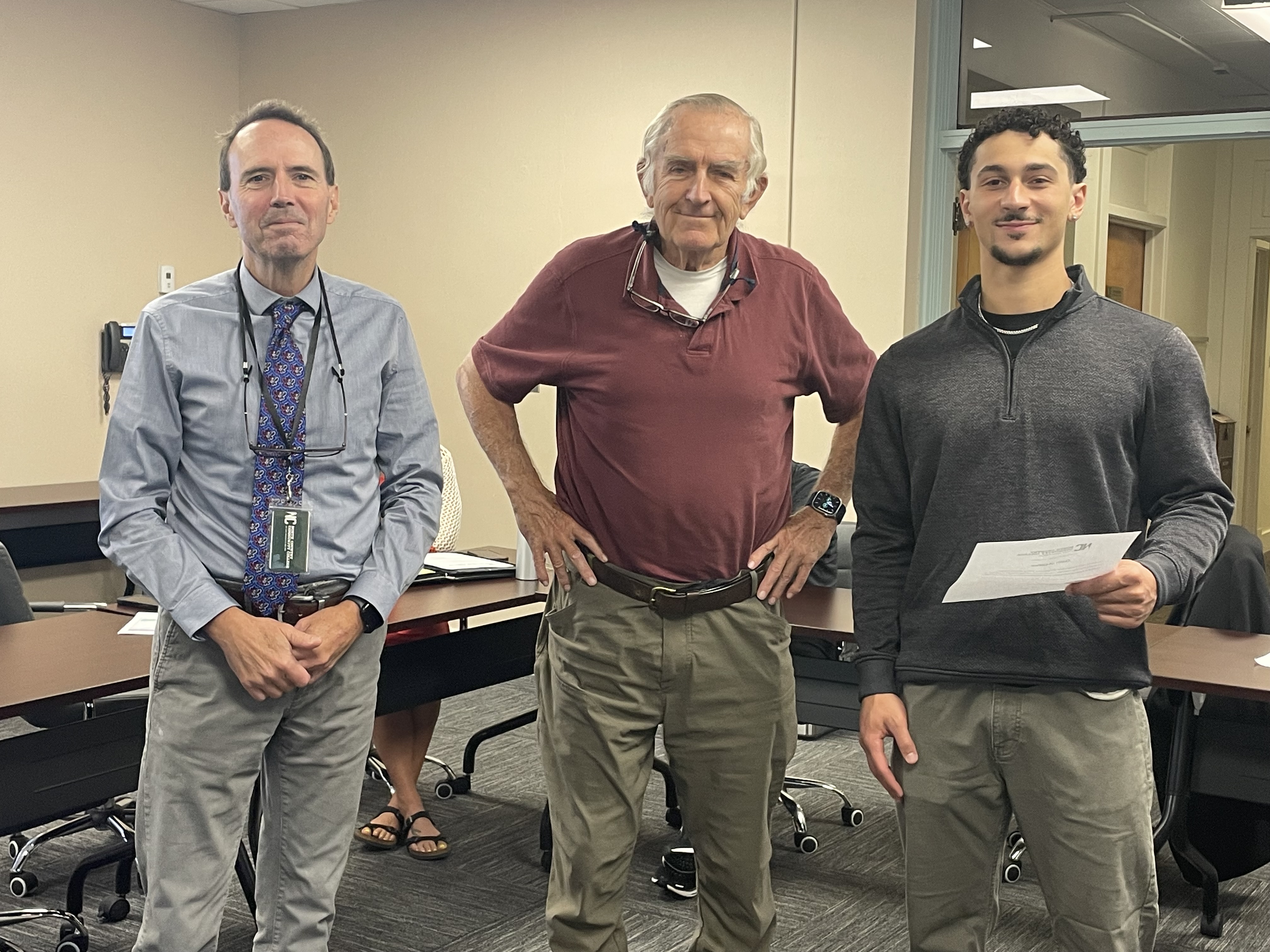 A college student stands in a classroom with two other people, facing the camera and smiling