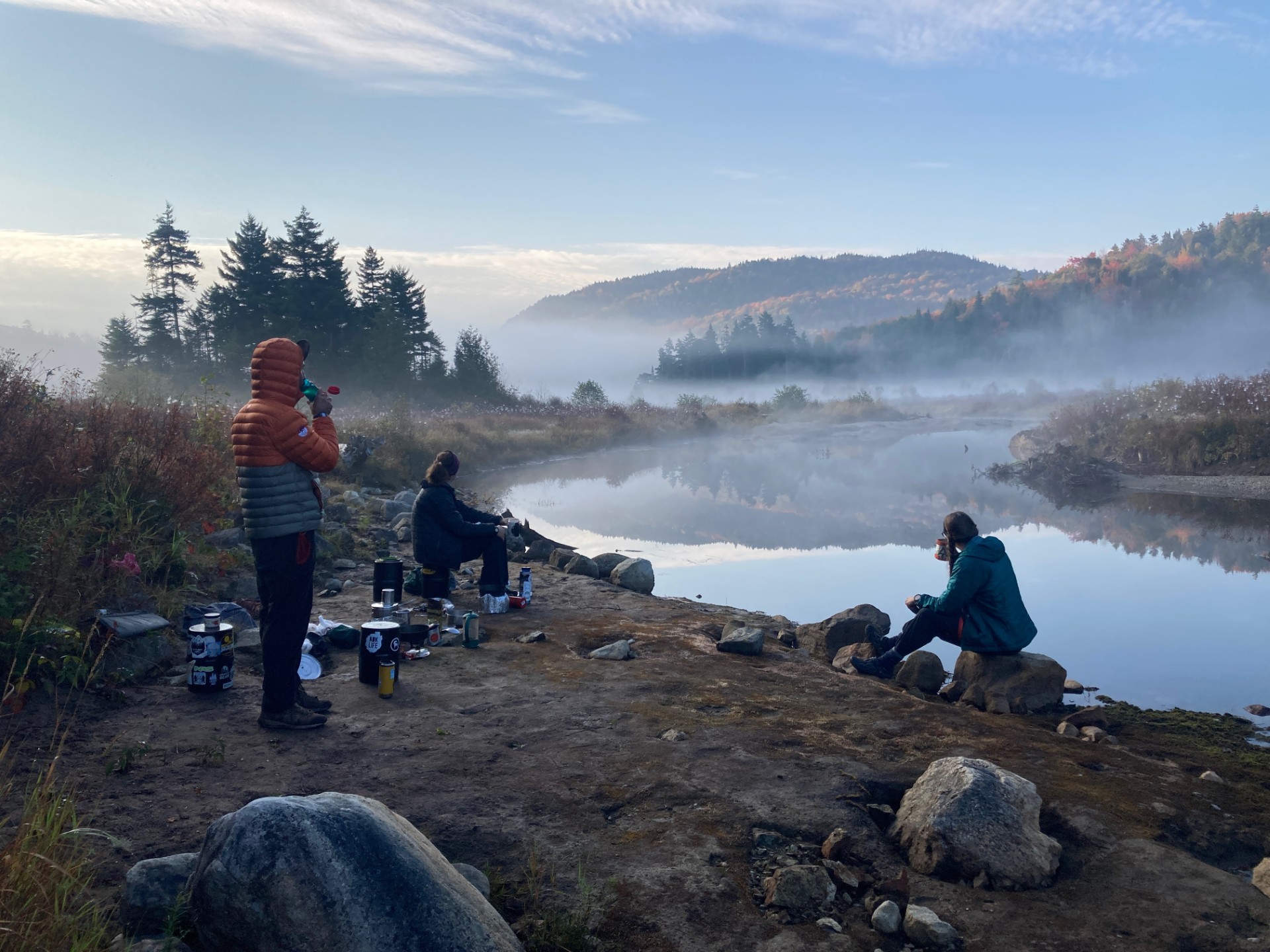 Students at a campsite along the water with fog covering the mountains in the distance