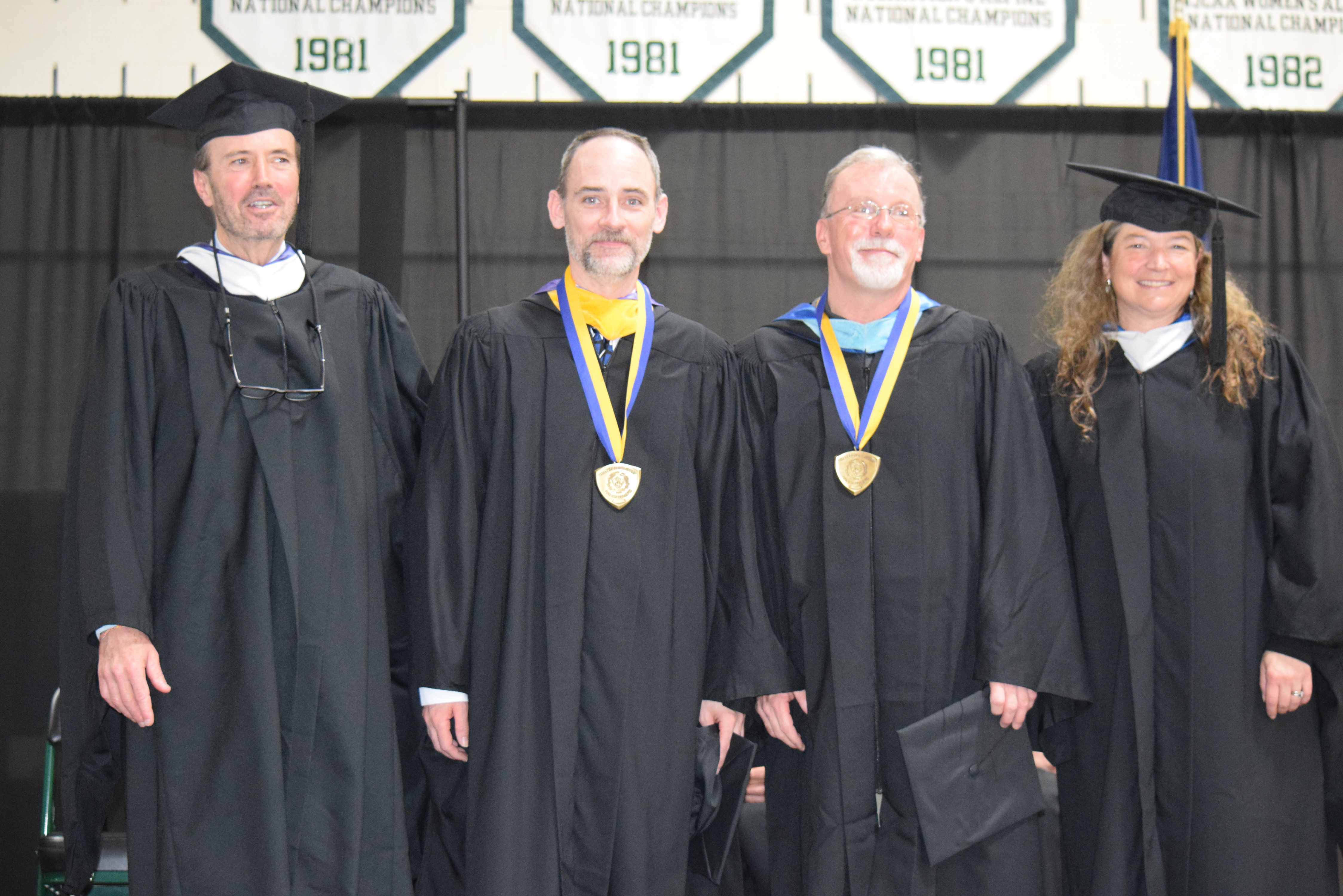 Four people stand on a stage in caps and gowns facing the camera and smiling.